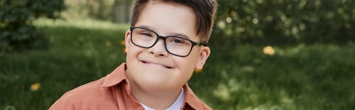 Preview: cheerful and genuine kid with down syndrome in eyeglasses smiling in park, portrait, banner