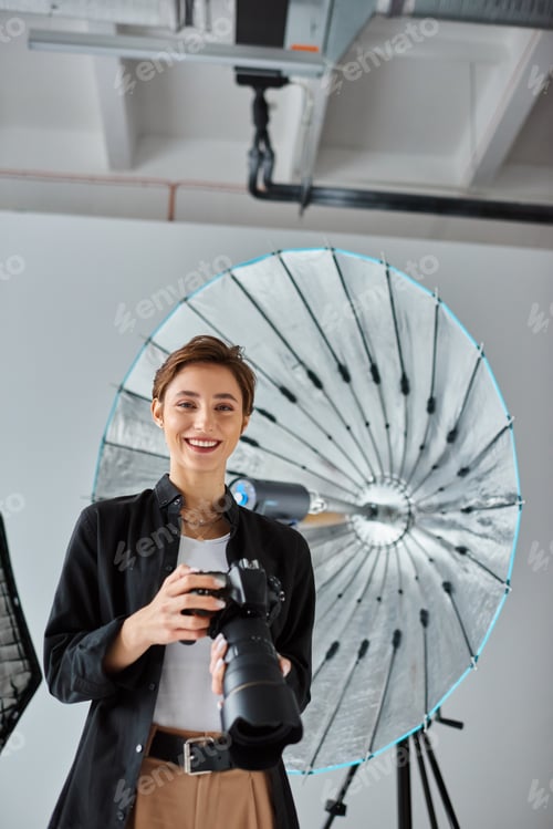 Preview: joyful short haired female photographer in casual attire posing with camera and smiling cheerfully