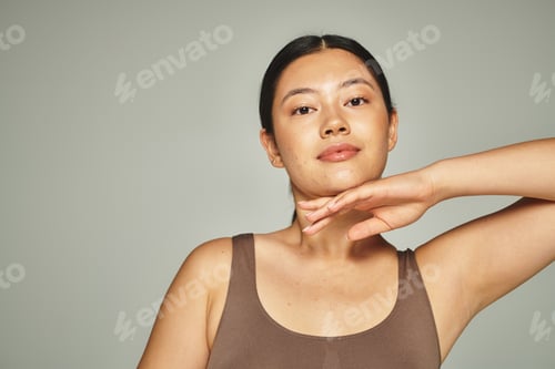 Preview: pretty asian woman in brown top touching her face on grey background, skin care and beauty