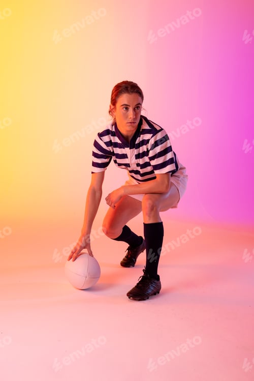 Preview: Caucasian female rugby player crouching with rugby ball over neon pink lighting
