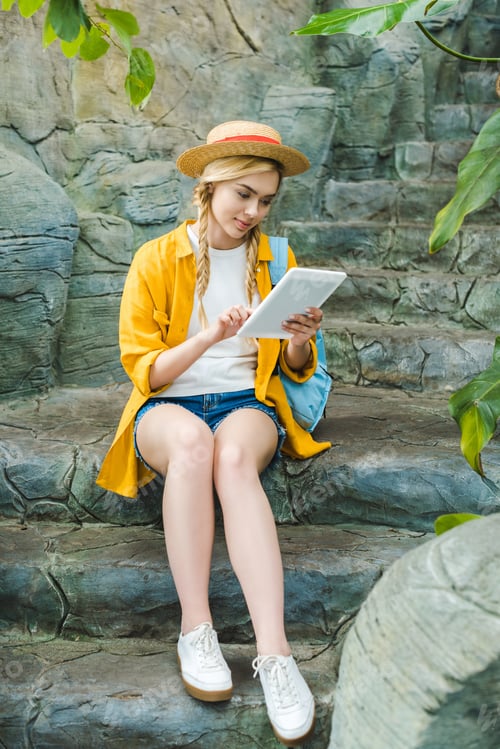 Preview: beautiful young woman in straw hat using tablet while sitting on stone staircase in park