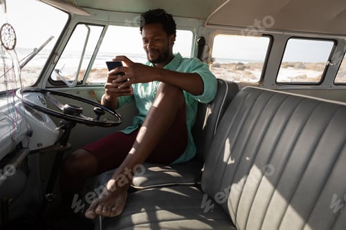 Preview: Front view of an African man sitting in a camper van while he is using his mobile phone at beach