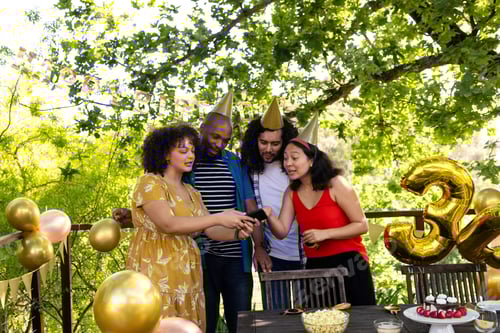Visualização: Amigos comemorando aniversário ao ar livre, usando chapéus de festa e olhando para o smartphone