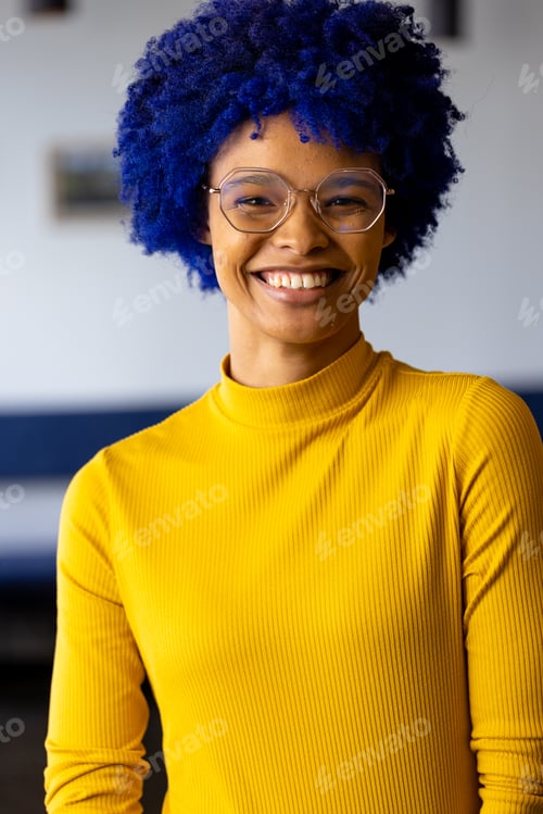 Preview: Portrait of happy biracial casual businesswoman with blue afro and glasses smiling in office