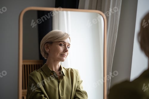Preview: Beautiful blonde middle aged woman standing in front of mirror smiling and enjoying own reflection.