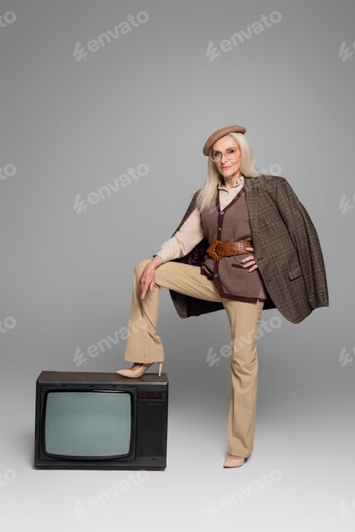 Preview: Elderly woman in heels and jacket standing near retro tv on grey background