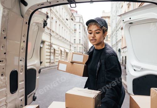Preview: Woman courier in uniform carefully unloading cardboard boxes from car for delivery