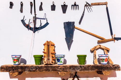 Preview: Old farm tools and colorful decorated terracotta pots, on a white wall in the sun.