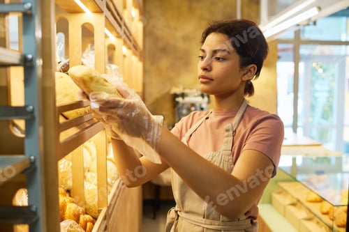 Preview: Young serious woman in beige cotton apron taking fresh bread from display
