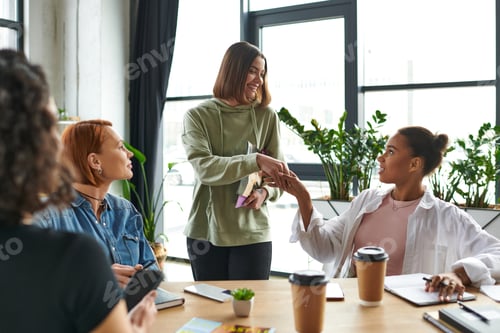 Preview: happy young woman holding magazines and shaking hands with african american member of interest club
