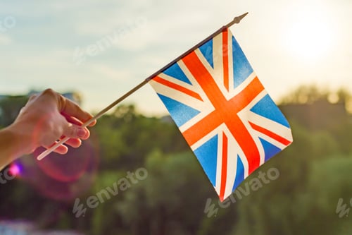 Preview: Hand holds Great Britainin flag an open window. Background blue sky, silhouette of the city, sunset