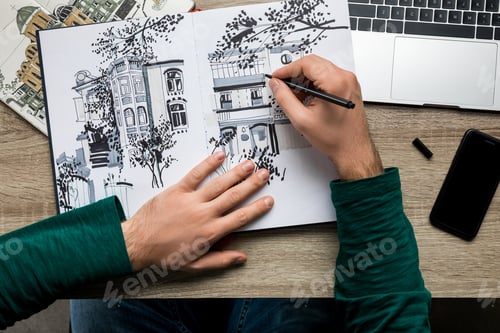 Preview: top view of mans hands drawing in album on wooden table next to smartphone and laptop