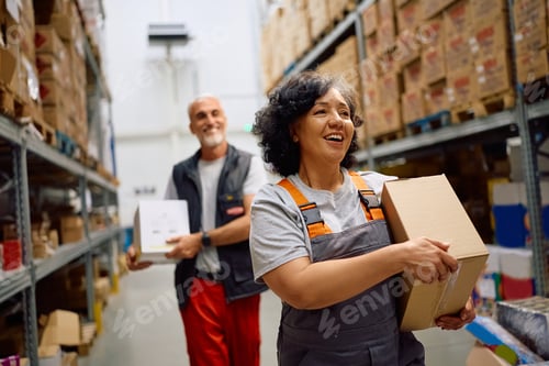 Preview: Happy worker and her colleague carrying cardboard boxes at distribution warehouse.