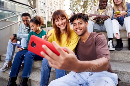 Preview: Multiracial group of young friends sitting on stairs hanging out, using mobile phones.