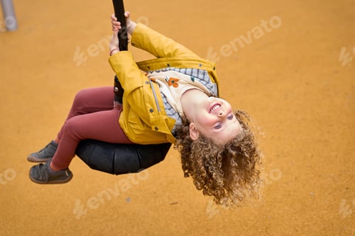 Preview: A young girl is hanging from a swing, wearing a yellow jacket and red pants
