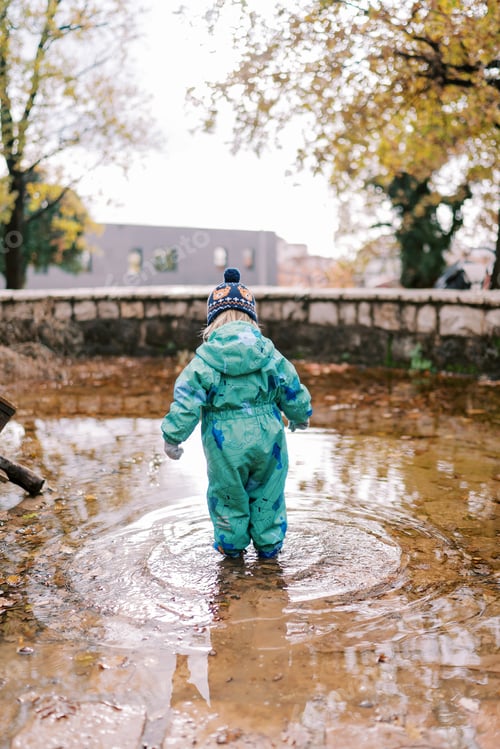 Preview: Little girl stands in a puddle and looks under her feet in the autumn park. Back view
