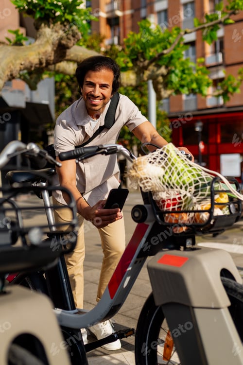 Preview: Young businessman using a smartphone next to a shared electric bicycle with a shopping basket