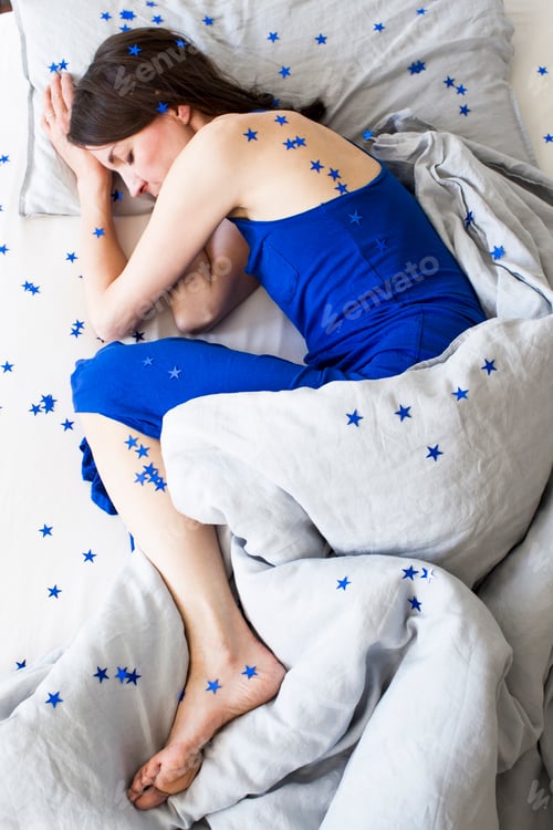 Preview: Overhead view of mature woman wearing blue pyjamas lying curled up in bed covered in stars