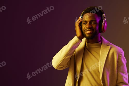 Preview: Young African man enjoying music with headphones in stylish beige blazer, smiling against a gradient