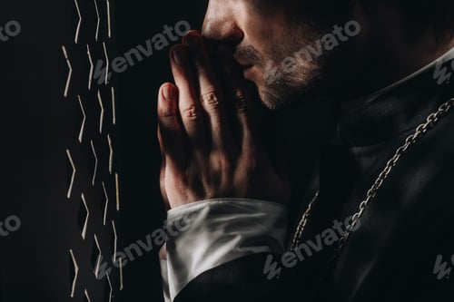 Preview: cropped view of catholic priest praying near confessional grille in dark with rays of light