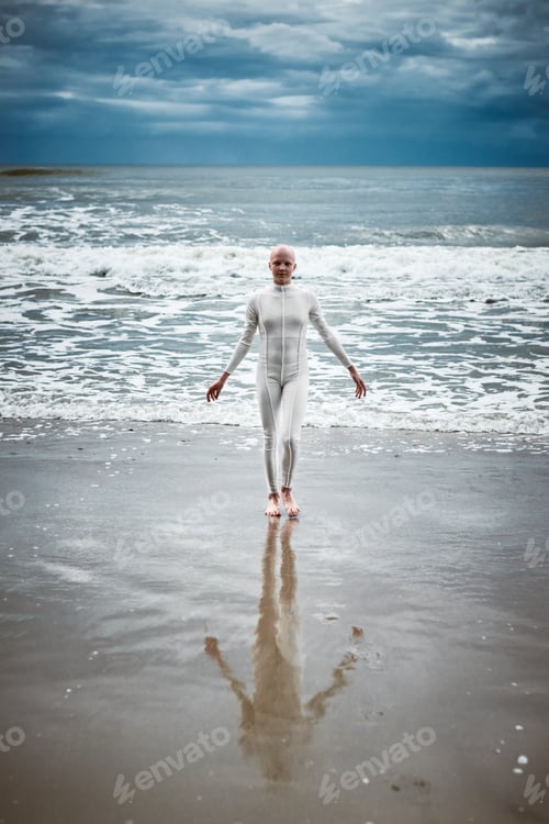 Preview: Hairless girl with alopecia in white futuristic suit comes out of cold restless sea on sandy beach