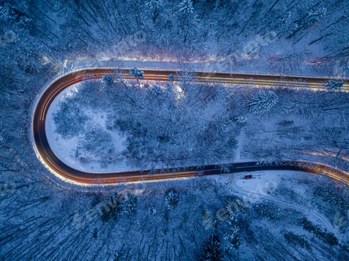 Preview: Car headlights illuminating a winding winter road and snowy fores.