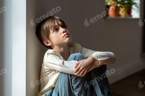 Preview: Lonely boy sadly looking up hugging knees sits near window at home alone. Kid psychological trauma