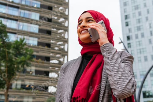 Preview: Delighted ethnic woman in hijab talking on smartphone in city