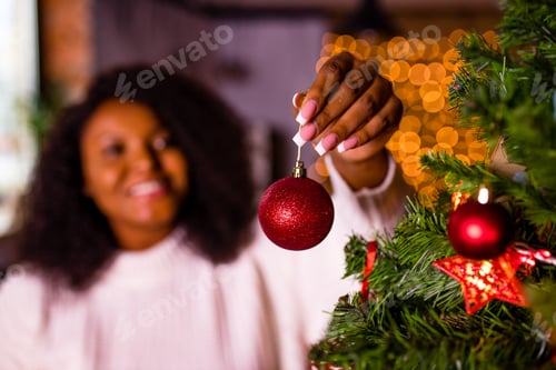 Preview: african woman in white sweater putting red ball on fir-tree at home