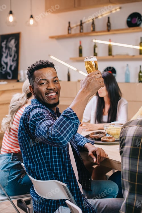Preview: cheerful african american man looking at camera while holding glass of beer near multicultural