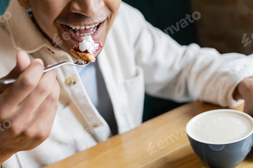 Preview: cropped view of happy african american man eating tasty cake near cup of cappuccino in coffee shop