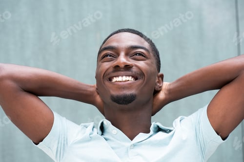Preview: Young African American man smiling with hands behind head against blue background and looking up