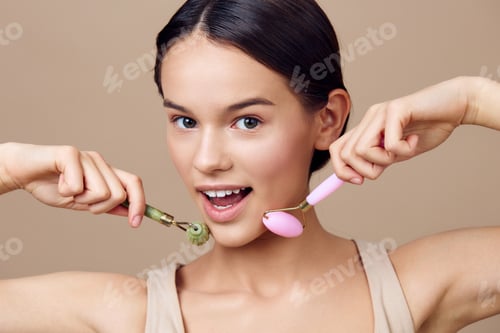 Preview: Young woman enjoying facial massage with green and pink rollers, showcasing a joyful expression