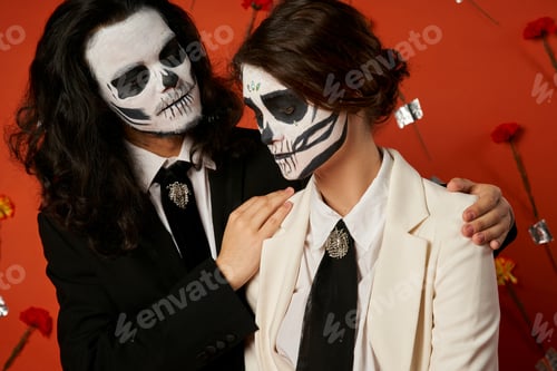 Preview: spooky man embracing shoulders of woman in suit, dia de los muertos couple on red floral backdrop