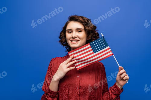 Preview: young cheerful student smiling at camera while holding USA flag isolated on blue
