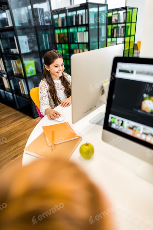 Preview: high angle view of adorable smiling schoolchild studying with computer in library