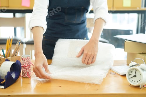 Preview: Asian female clothes shop owner folding a t-shirt and packing in a cardboard parcel box.