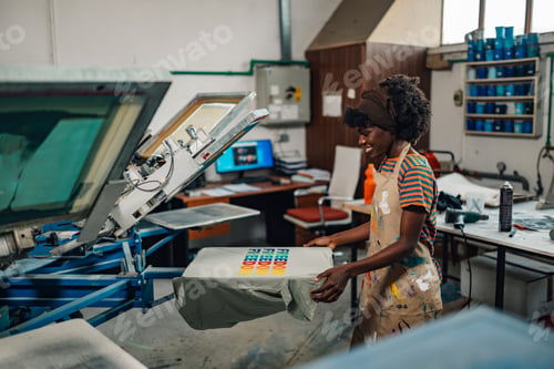 Preview: Happy diverse graphic worker taking screen printed shirt of machine.