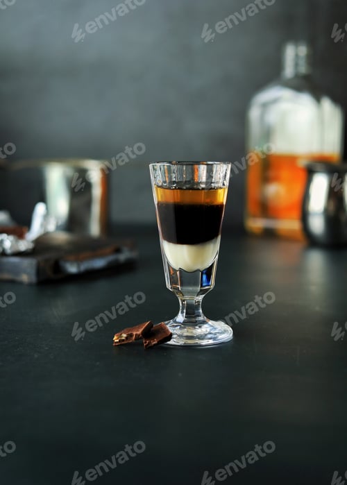 Preview: A three-layer cocktail shot with whiskey and liqueurs in a glass and chocolate next to it on a dark