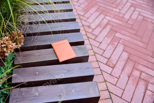 Preview: Orange passport lost on wooden bench beside brick pavement and Greenery