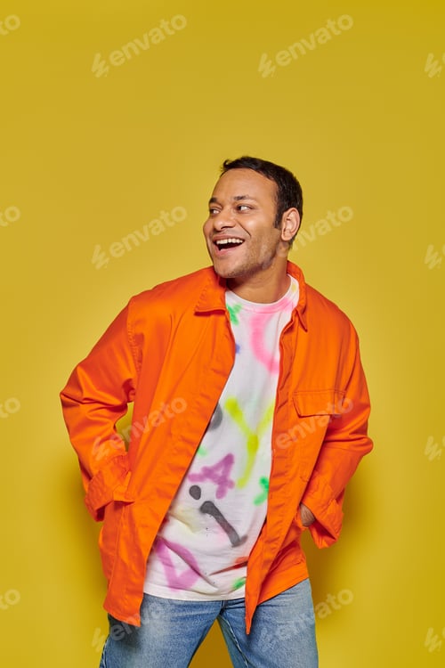 Preview: portrait of joyful indian man in orange jacket posing with hands in pockets on yellow backdrop