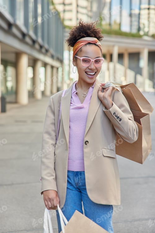 Preview: Happy young woman enjoys holiday discounts in shopping mall walks at street with paper bags wears tr