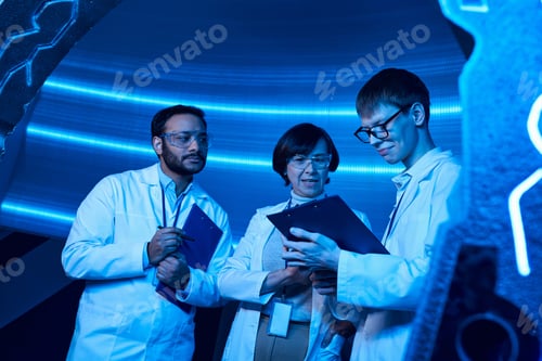 Preview: young intern looking at clipboard near multiethnic colleagues in neon-lit discovery center
