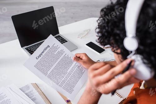 Preview: Devices near blurred indian freelancer in headphones working with paper with copywriting lettering