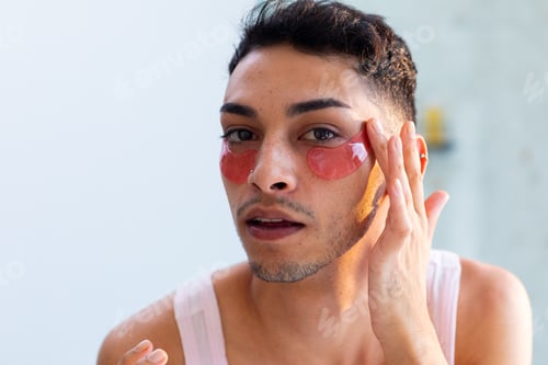 Preview: Biracial transgender man looking in mirror and applying under-eye patches in bathroom