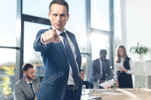 Preview: serious businessman pointing at camera in conference hall with blurred colleagues on background