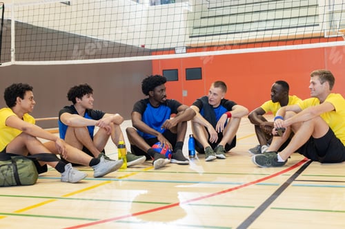 Preview: Diverse male partners sitting on gym court under net with volleyball, water bottles and sports bag