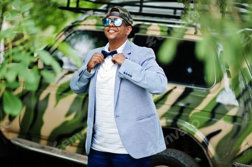 Preview: Stylish arabian man at sunglasses, cap and bow tie posed against military car. Arab model boy.