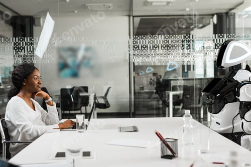 Preview: smiling african american businesswoman and robot sitting at desk in conference hall