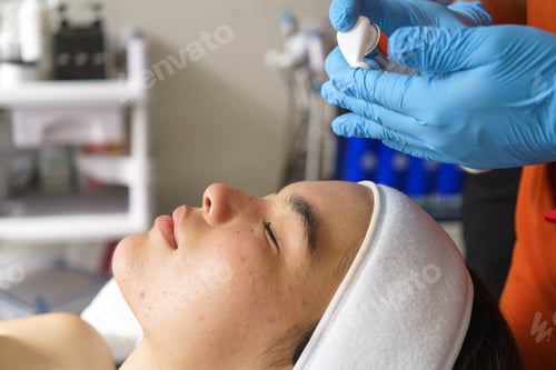 Preview: Beautician applying product on face of asian young woman with acne